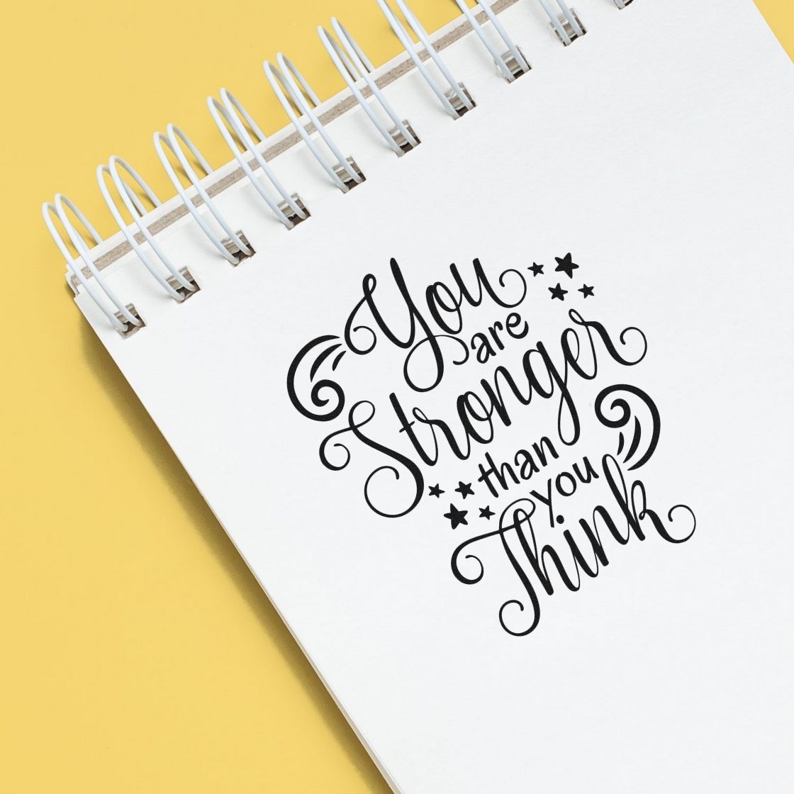 Black motivational stamp design on white spiral notebook with yellow background, reading You are stronger than you think.