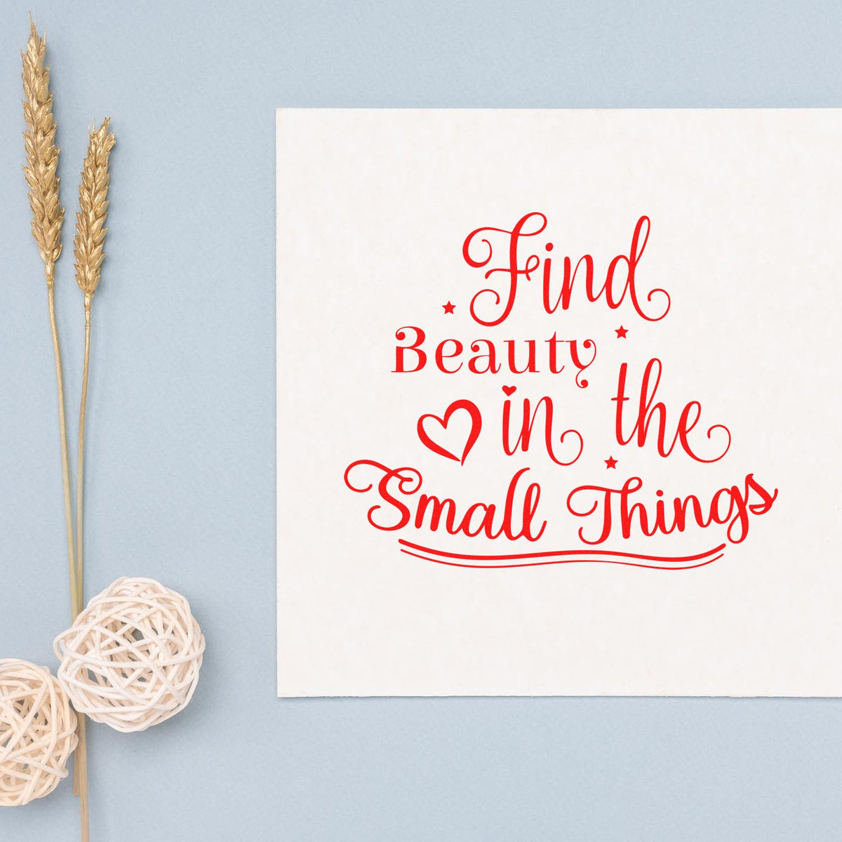 Red custom stamp and seal reading Find Beauty in the Small Things on a white card, with blue background and dried wheat.