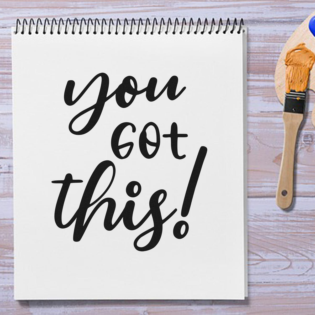 Black you got this! script on a white spiral notebook beside a gold-bristled paintbrush, custom stamp and seal design.