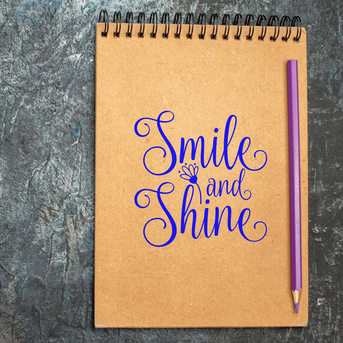 Blue script stamp and seal reading Smile and Shine on a tan spiral notebook with black coil and purple pencil.