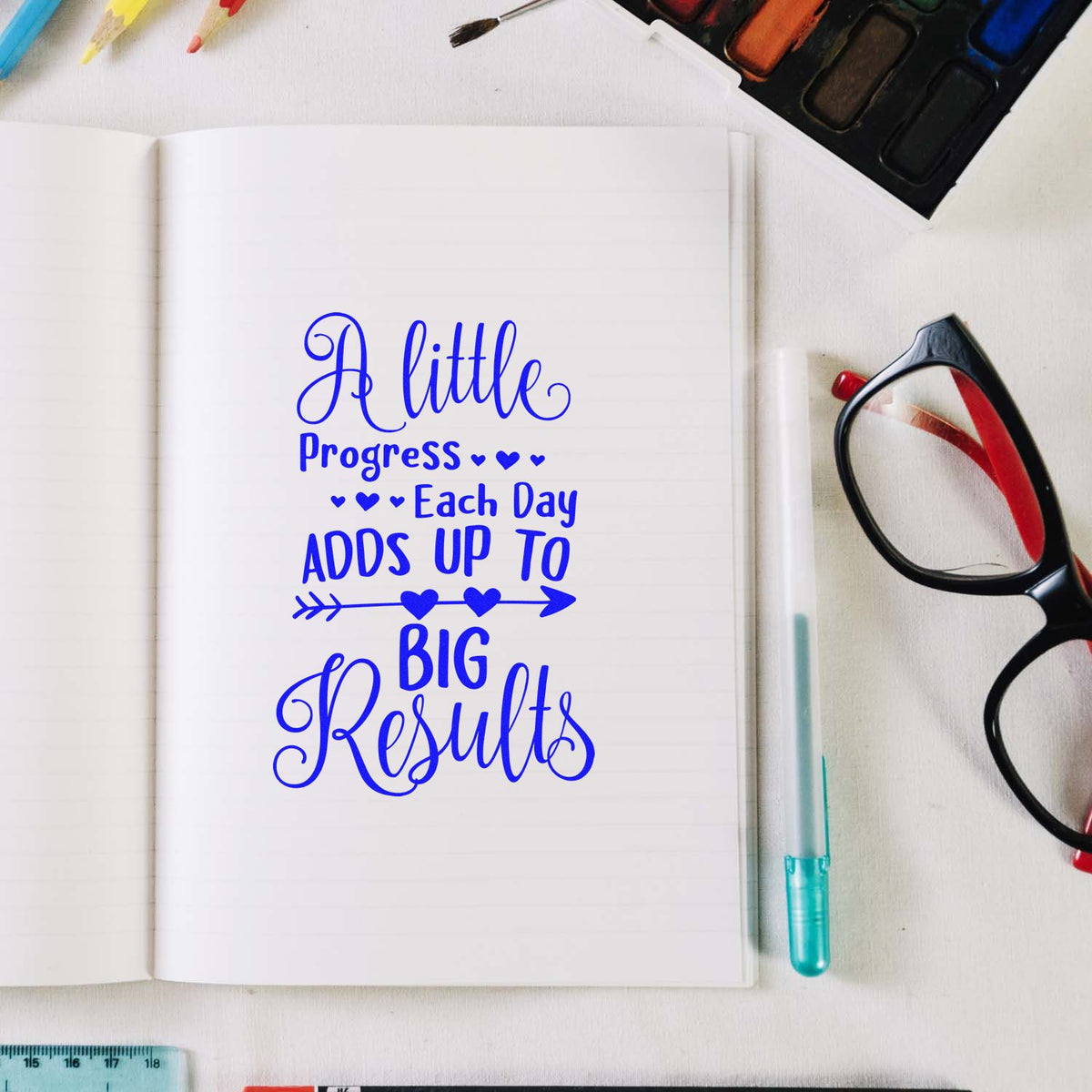 Blue self-inking stamp and seal design in notebook reading A little progress each day adds up to big results, with art supplies.