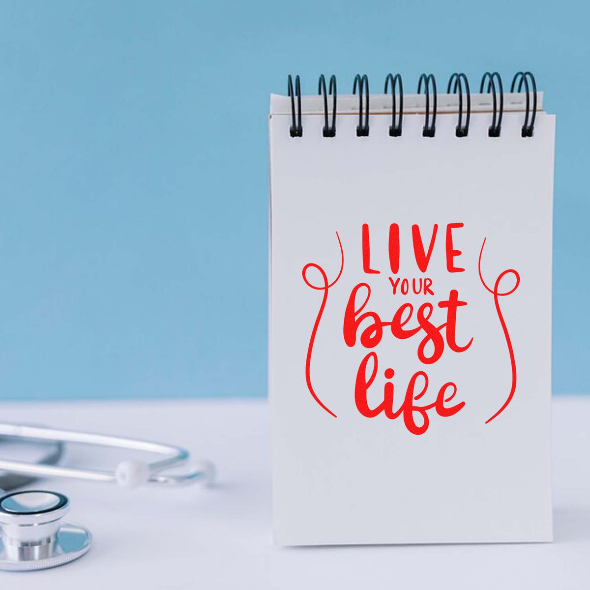 Red custom stamp reading Live your best life on a spiral notebook, with a stethoscope on a light blue background.
