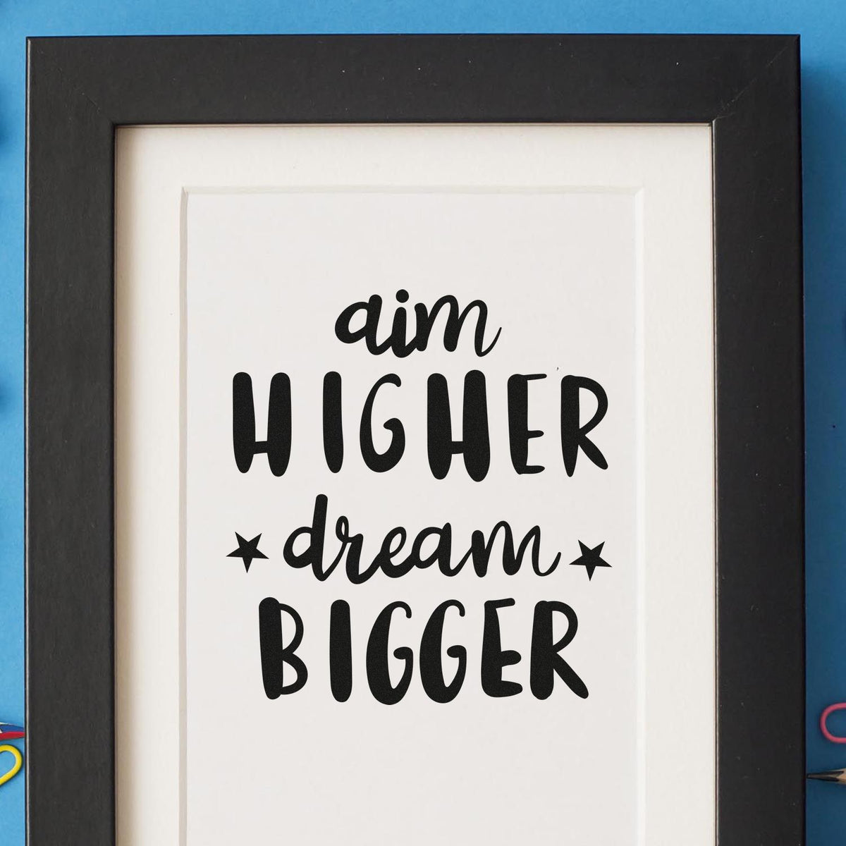 Black stamp and seal artwork in a black frame on blue background, reading aim higher dream bigger.