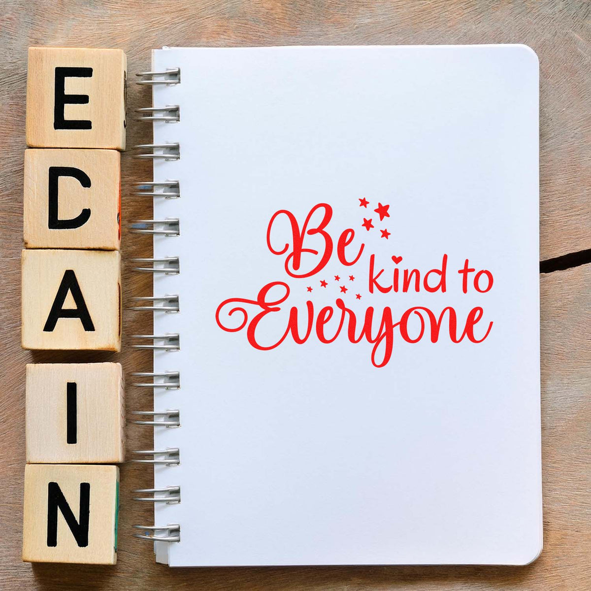 Wooden letter stamp blocks beside a spiral notebook stamped in red with Be kind to Everyone and star accents.