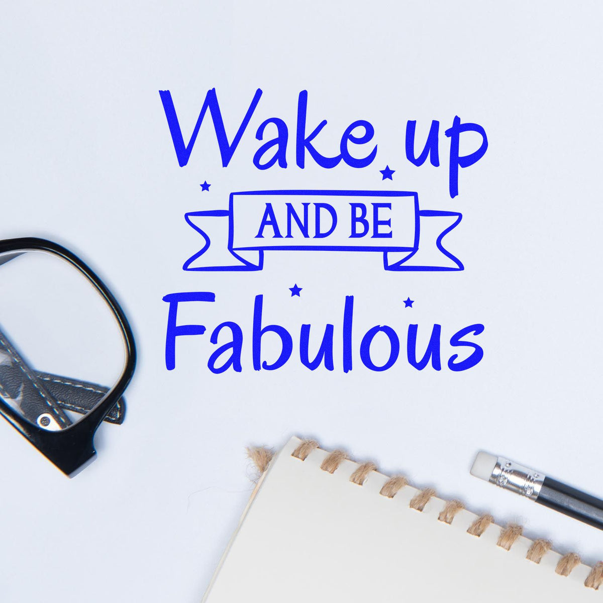 Blue stamp-and-seal design reading Wake up and be Fabulous beside a stamp tool, notebook, and pencil on white background.