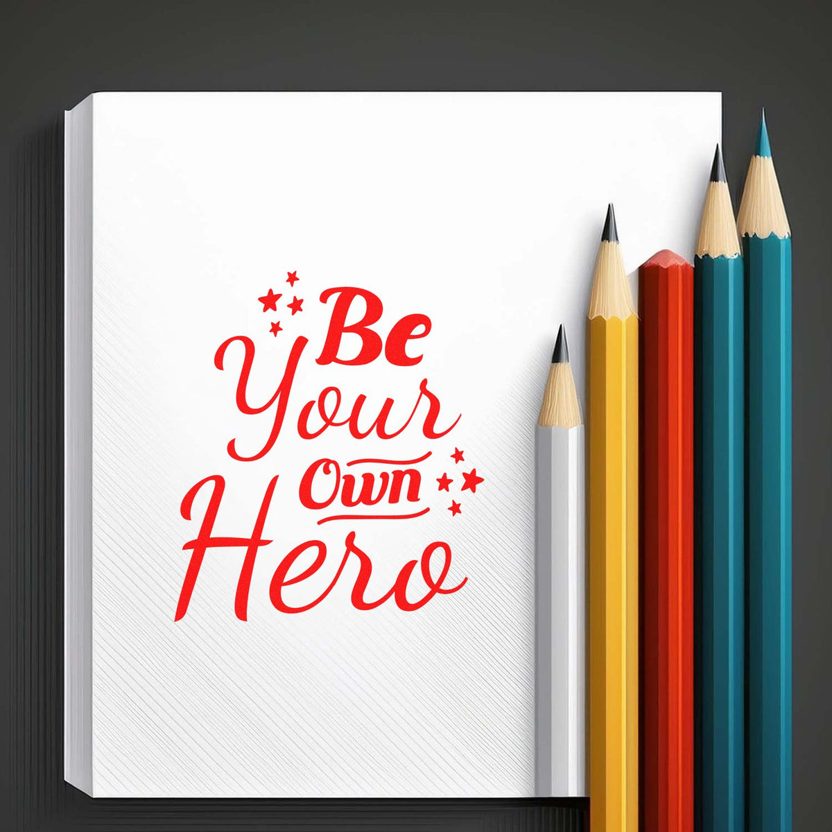 White stamp and seal pad with red Be Your Own Hero text, beside colorful pencils on a dark background.
