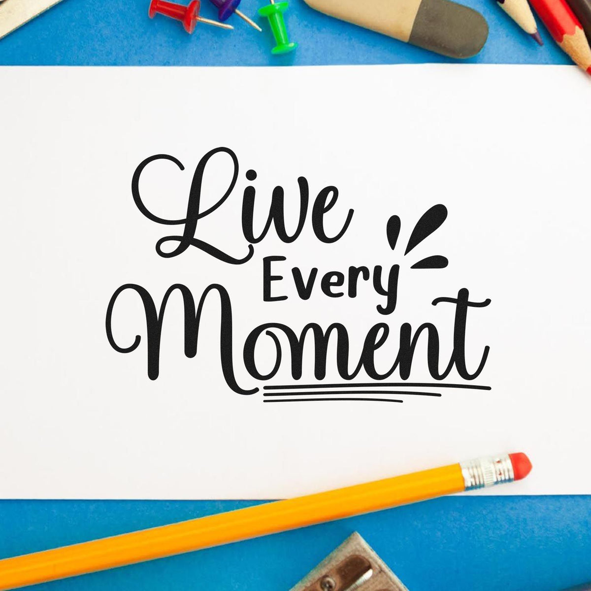 Black custom stamp and seal reading Live Every Moment on white paper, with colorful pencils on blue.