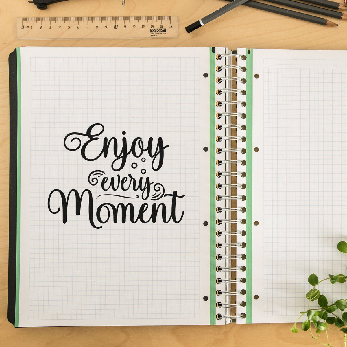 Black stamp and seal design reading Enjoy every Moment on white grid notebook pages with green spiral binding.