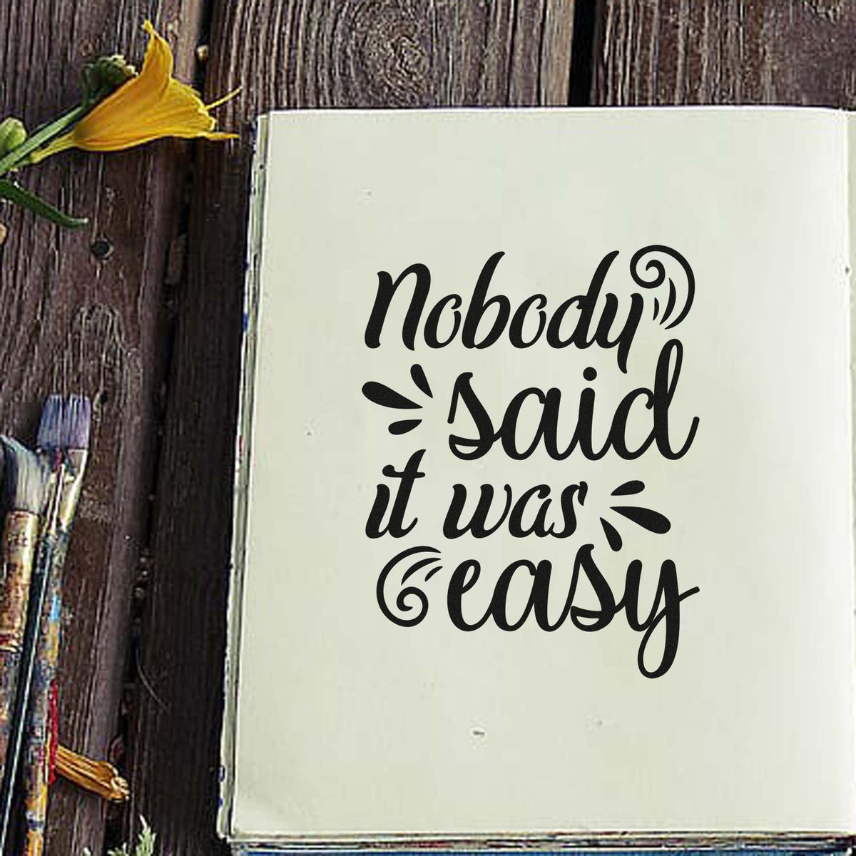 Black script stamp and seal reading Nobody Said it was easy on off-white paper, with decorative swirls on rustic wood and yellow flower.