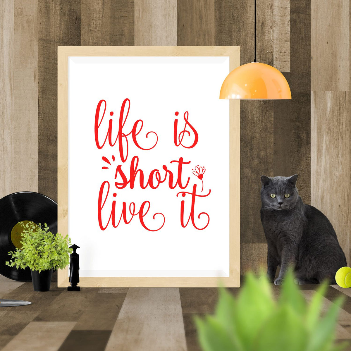 Red calligraphy stamp and seal design reading life is short live it in a light wood frame against rustic wood paneling.