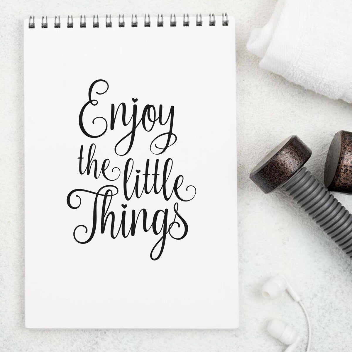 White spiral notebook with black Enjoy the little Things design, beside metallic stamp and seal tools on a white background.