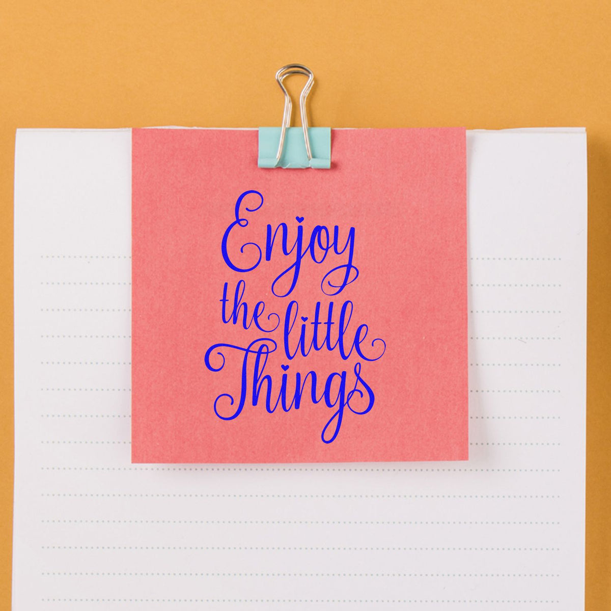 Coral stamp-and-seal sample with blue Enjoy the little things script, clipped to a white dotted notebook page on an orange background.