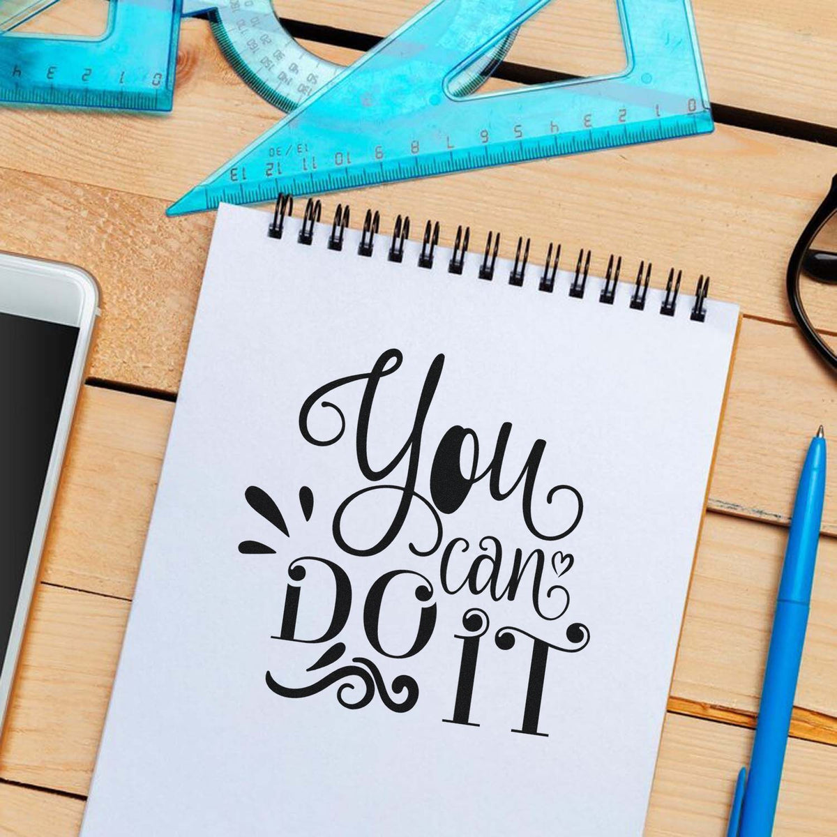 Black decorative stamp and seal reading You can do it on a white notepad, with blue rulers and pen on a wood desk
