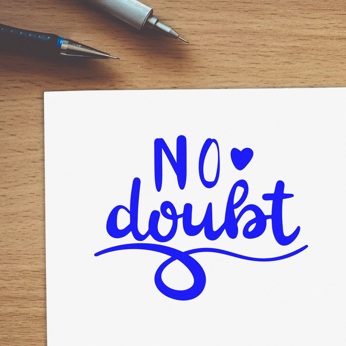 Blue No doubt stamp-style seal with decorative flourish on white paper, on a wooden desk beside two pens.