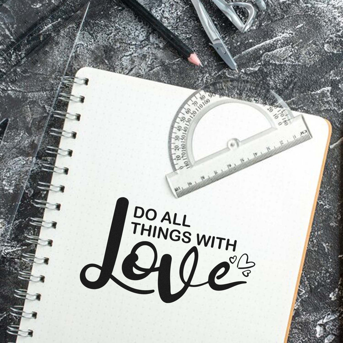 Black Do All Things With Love stamp on a white spiral notebook, with clear ruler and protractor on a dark textured background.