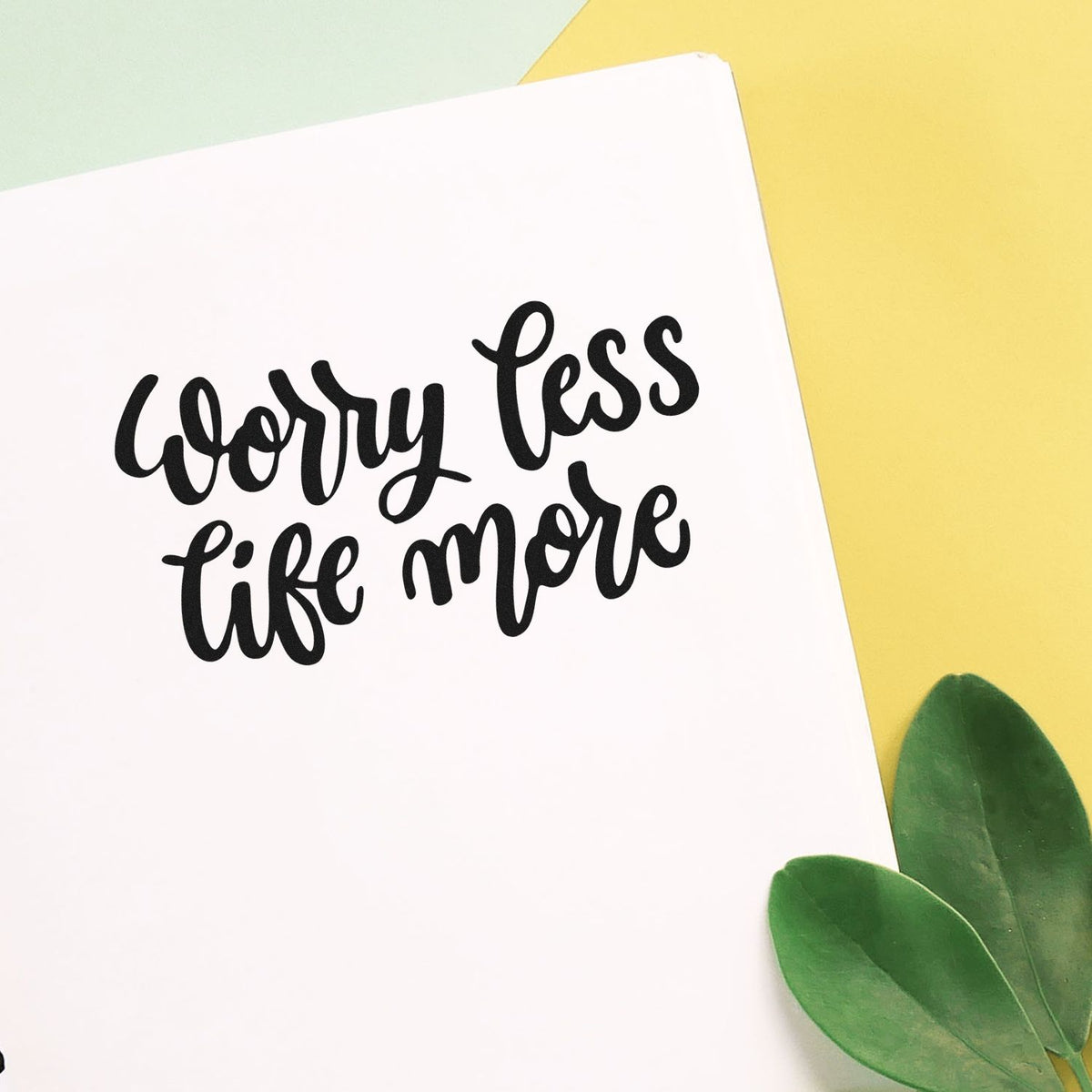 Custom black script stamp reading worry less life more on white card, with pastel yellow background and green leaves.