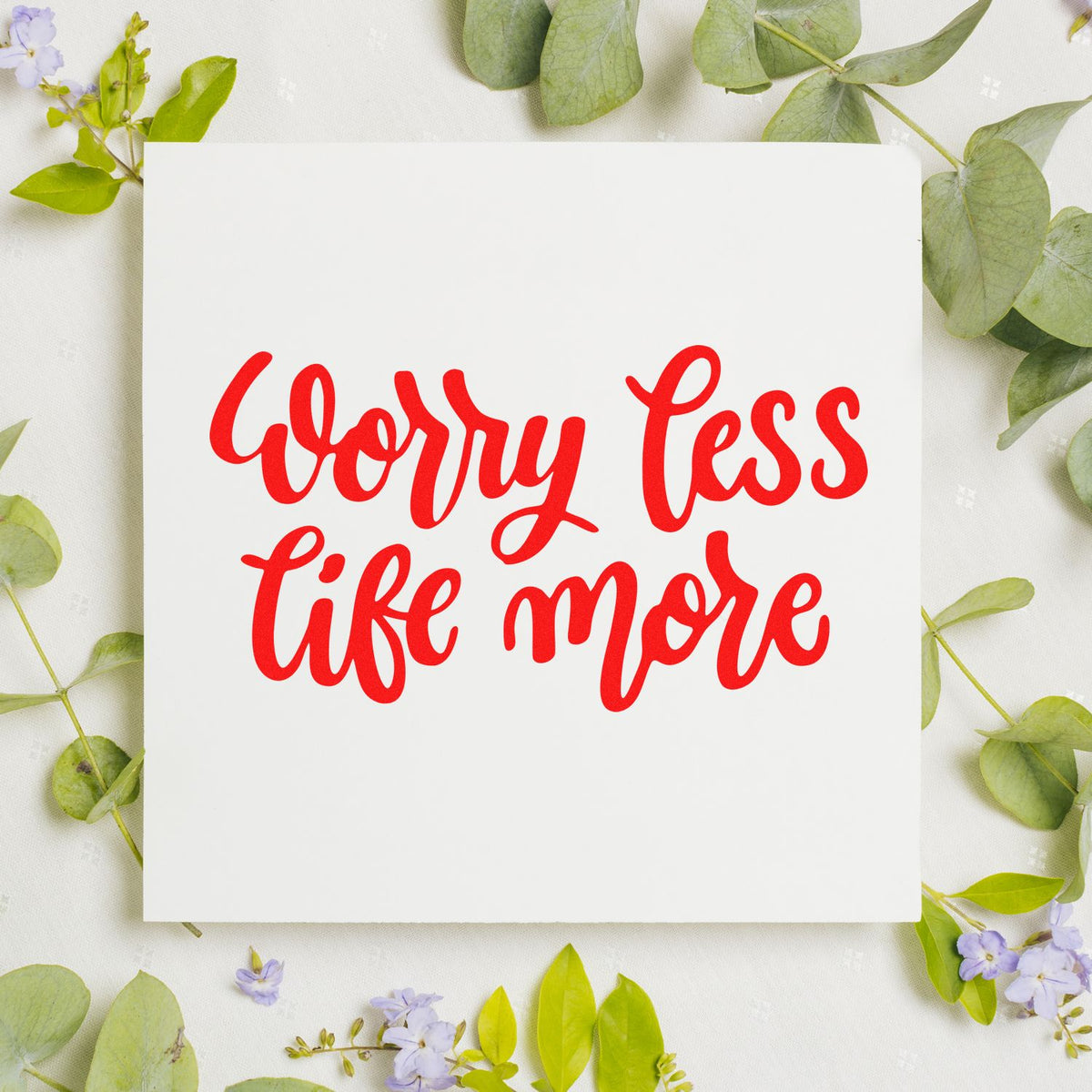 Red Worry Less Life More stamp/seal on white square, framed by green leaves and small purple flowers.