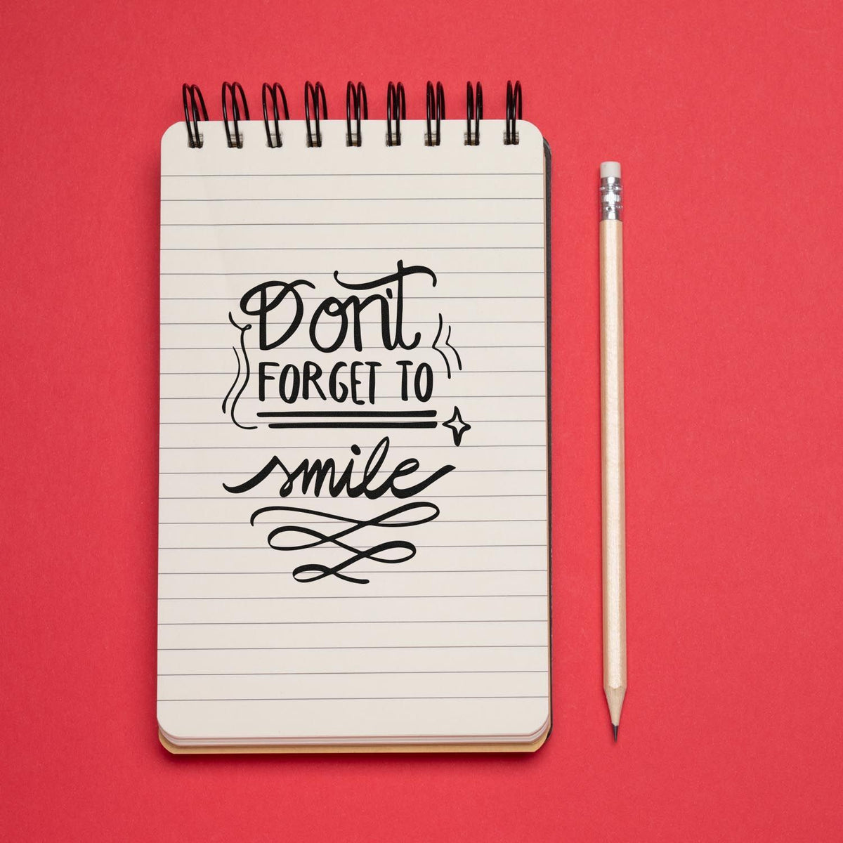 Black Dont forget to smile stamp design on a lined spiral notebook, with pencil, against a bright red background.