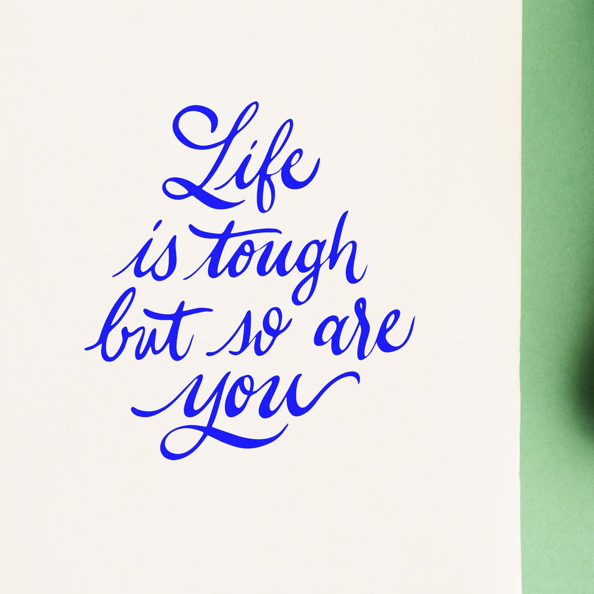 Blue script stamp reading Life is tough but so are you on white seal paper with a green edge.