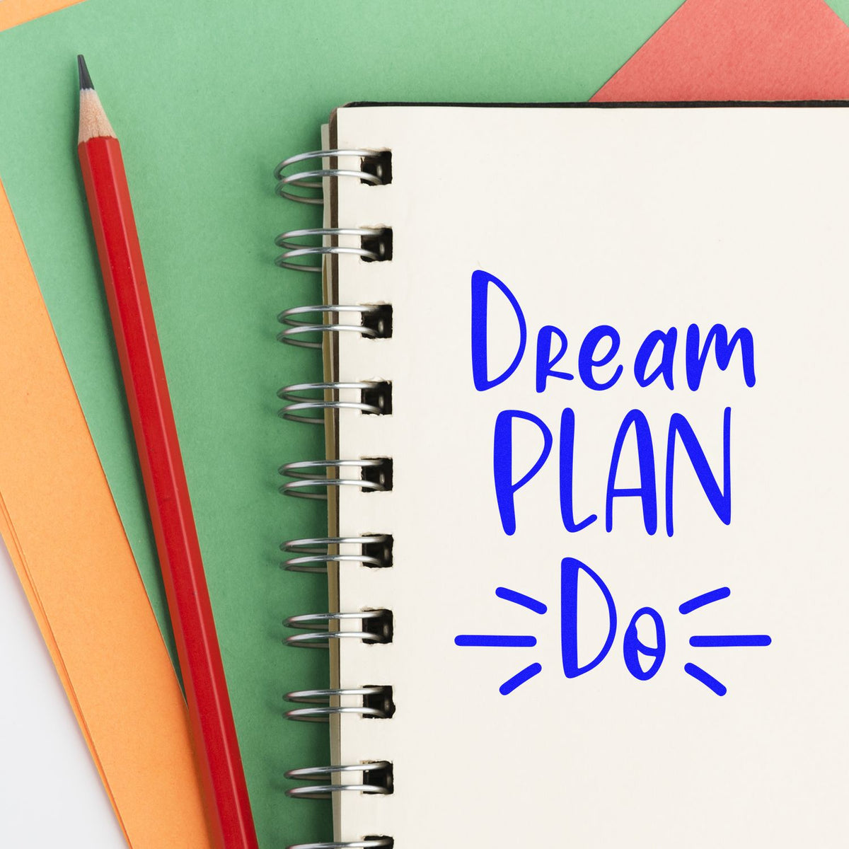 Blue Dream Plan Do stamp on a spiral notebook, with a red pencil and colorful paper background.