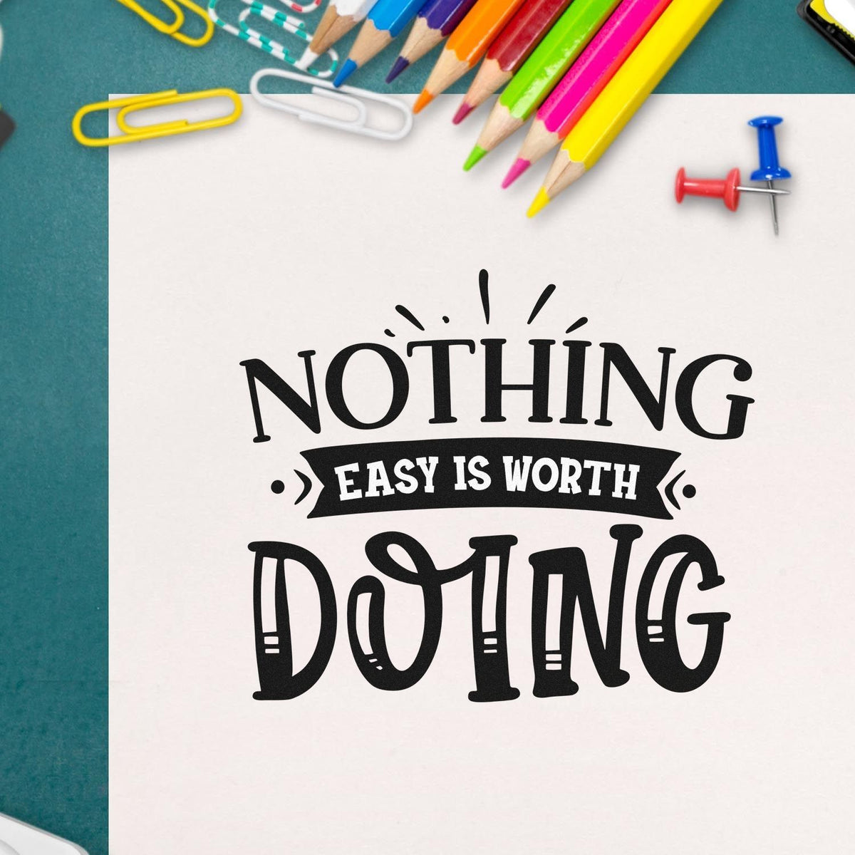 Black Nothing Easy Is Worth Doing stamp and seal design on white paper, with colorful pencils, clips, and push pins.
