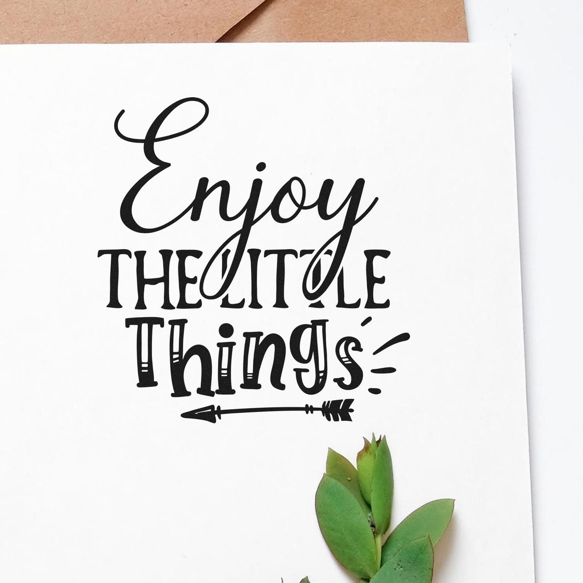 Black rubber stamp seal with Enjoy the Little Things in script on white paper, accented by a small green sprig.