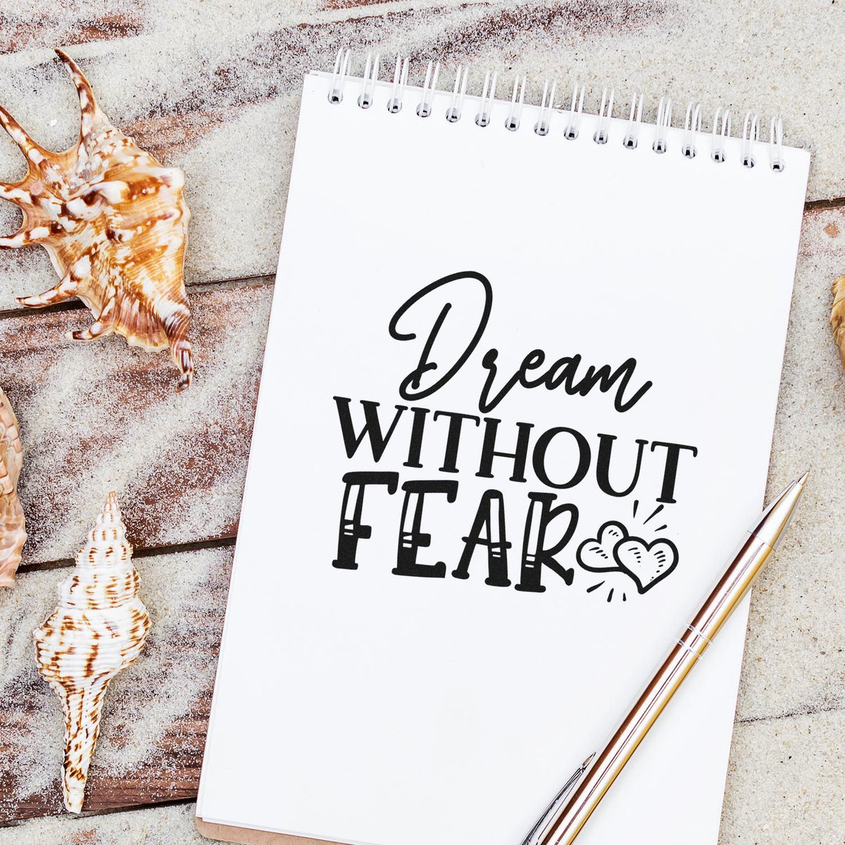 White spiral notebook with black Dream Without Fear stamp design, heart icons, seashells, and gold pen on sandy beige background.