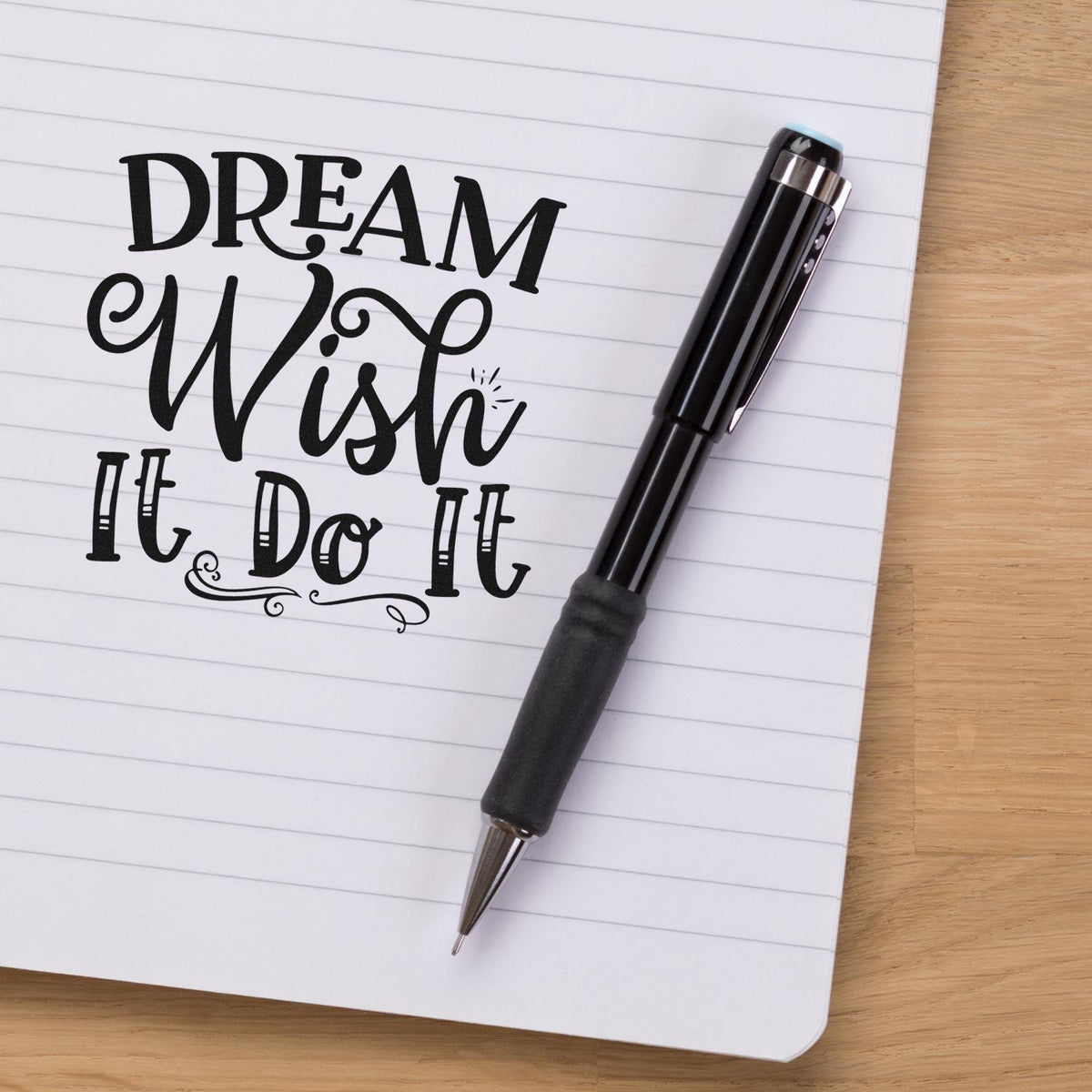 Black self-inking stamp and seal pen beside lined notebook with Dream Wish It Do It text on white paper.