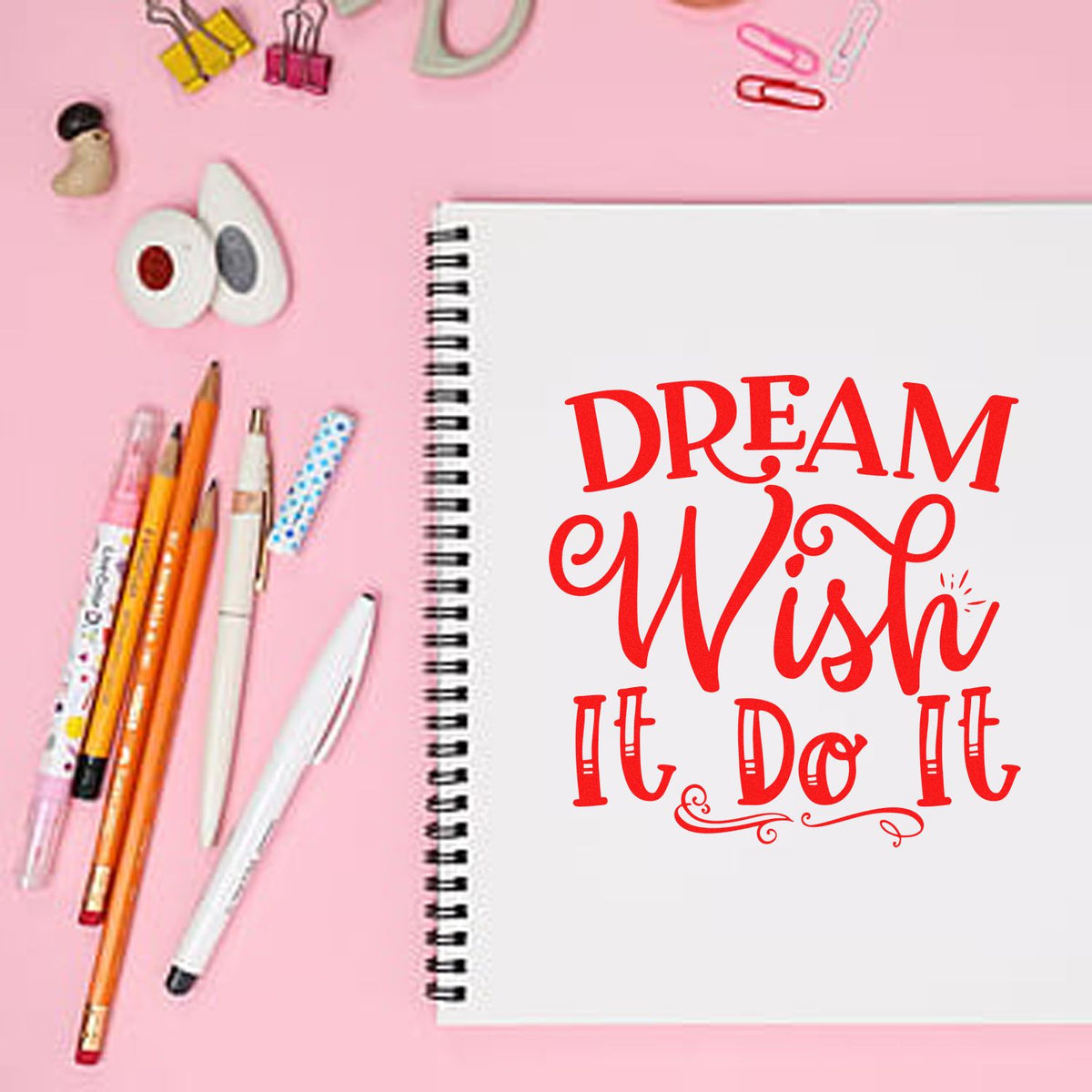 Pink flat lay of stamp and seal products with a spiral notebook stamped Dream Wish It Do It, plus pens and clips.