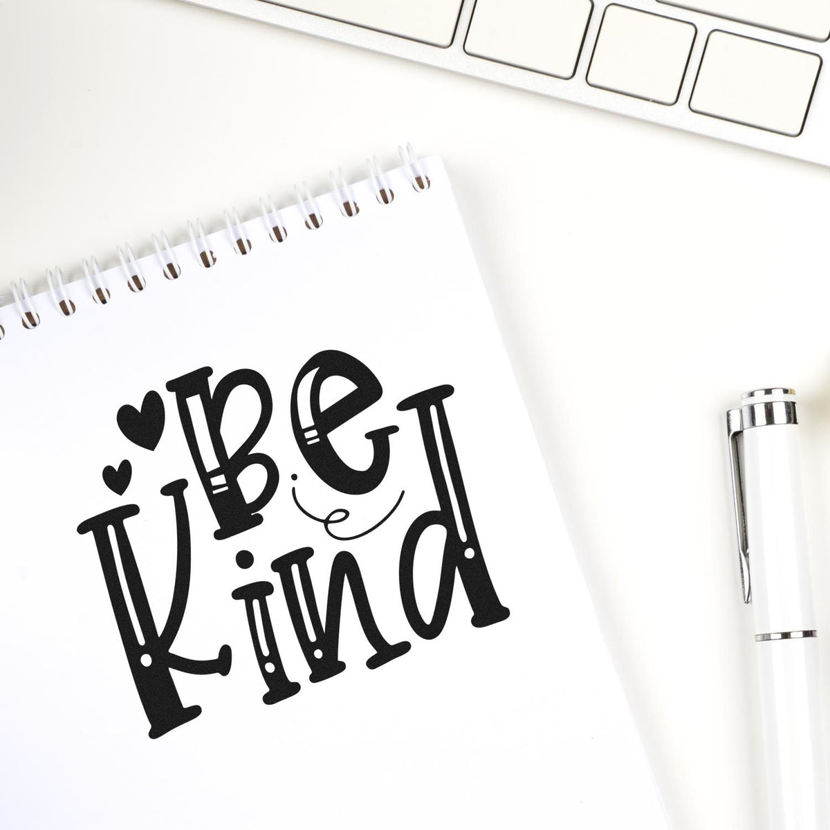 Black Be Kind stamp design on a white spiral notebook, with a white pen and keyboard on a clean desk.