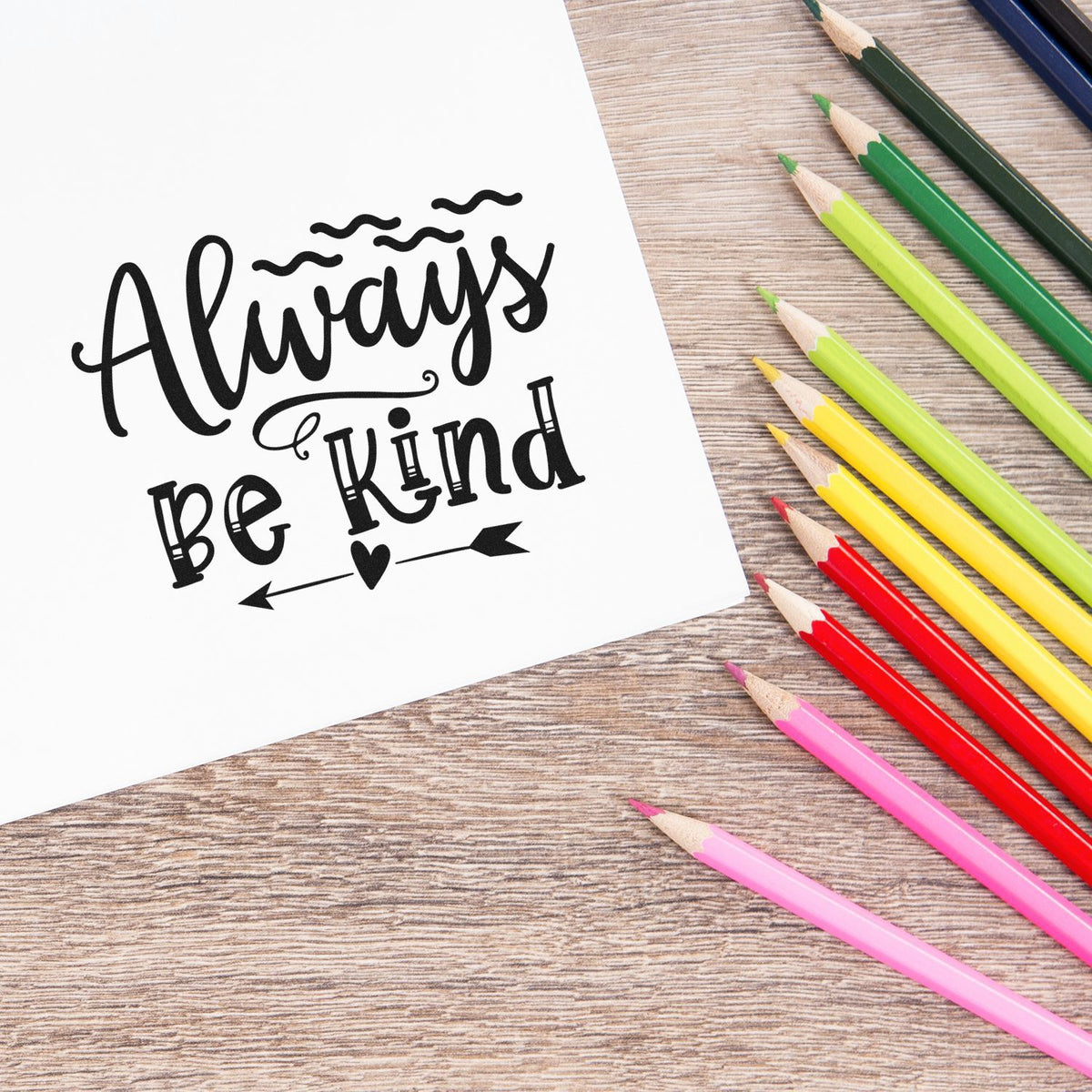 Custom stamp and seal design reading Always Be Kind in black, with colorful pencils on a wood desk.
