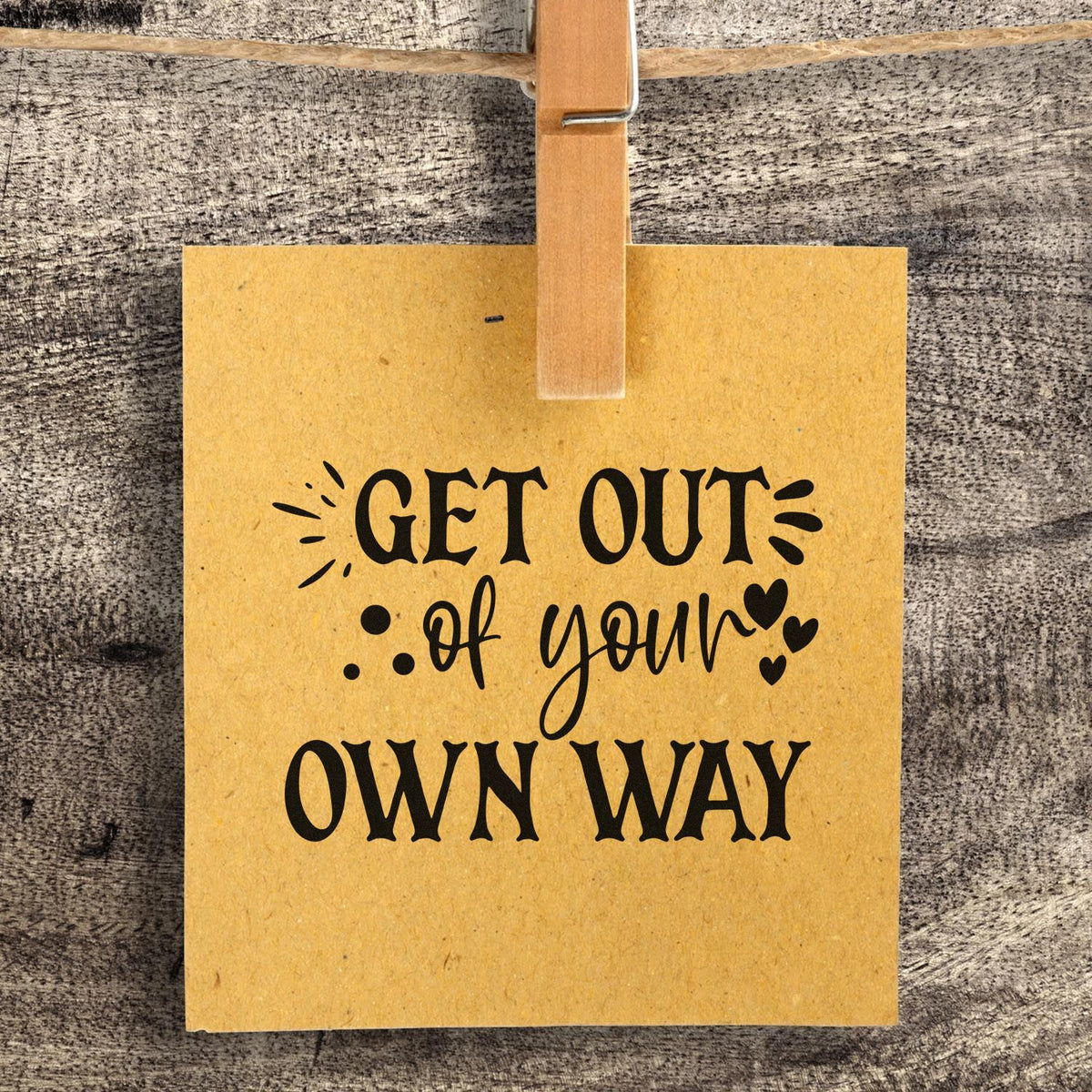 Yellow kraft paper stamp seal hanging on rustic wood, with black Get Out of Your Own Way lettering and small hearts.