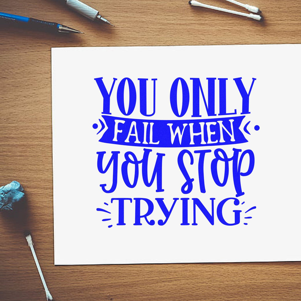 Blue stamp/seal design on white paper reads You only fail when you stop trying in bold blue on a wood desk with tools.