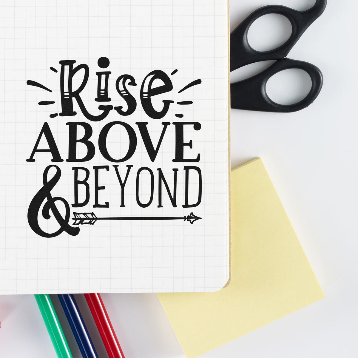 Black Rise Above & Beyond stamp and seal design on graph paper notebook, with scissors, sticky note, and colored pens.