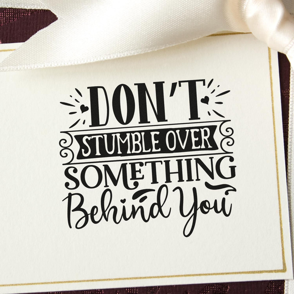 Black decorative stamp seal on white card with ribbon, reading Dont stumble over something behind you.