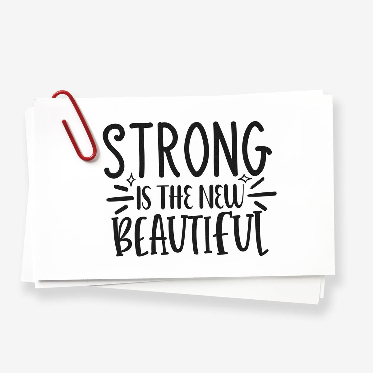 White stamp and seal card with black Strong is the new beautiful text and a red paperclip on layered white sheets.