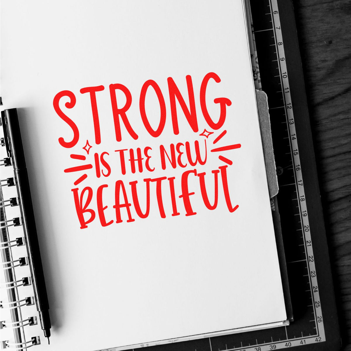 Red stamp/seal design reading Strong is the New Beautiful on a white notebook page with black pen and ruler.