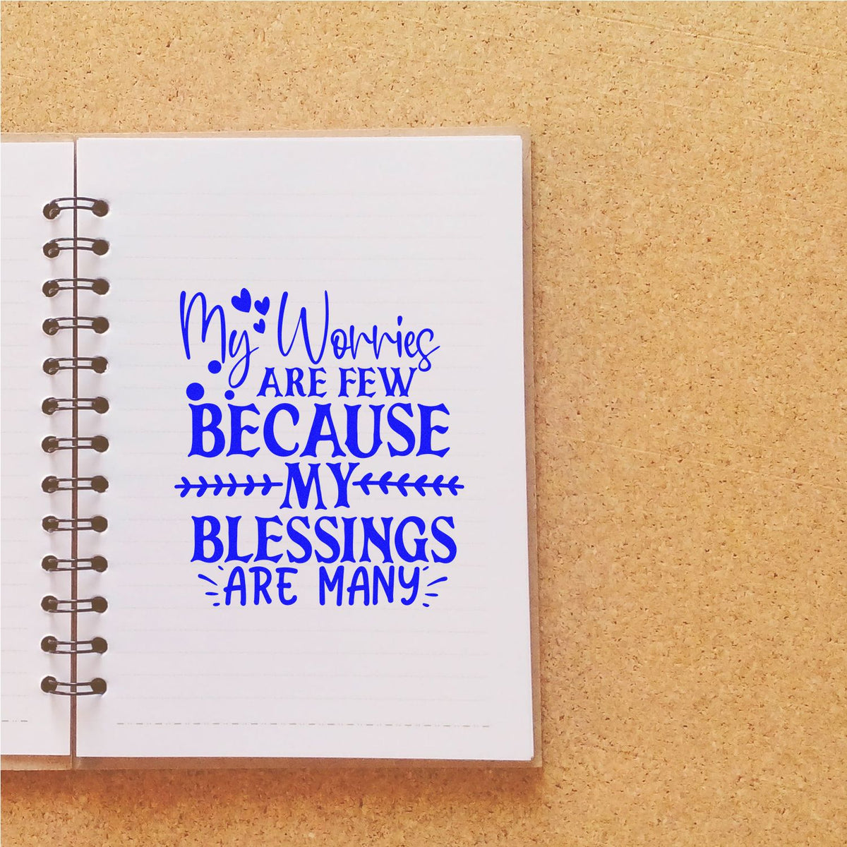 Blue blessing quote stamp on white spiral notebook page, reading My worries are few because my blessings are many, on tan background.