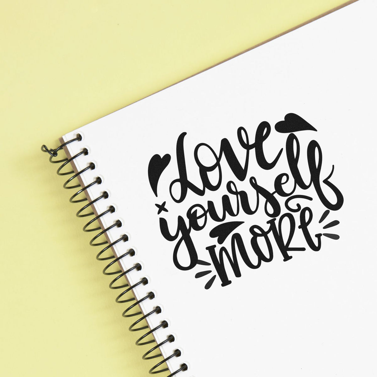 Black love yourself more stamp and seal design on a white spiral notebook with a pale yellow background.