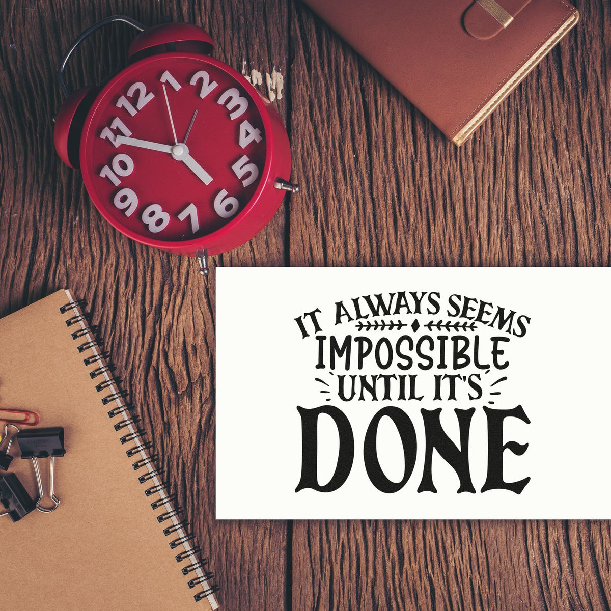 Red alarm clock beside a white motivational rubber stamp seal reading Done on a rustic wood desk, with notebook and leather planner.