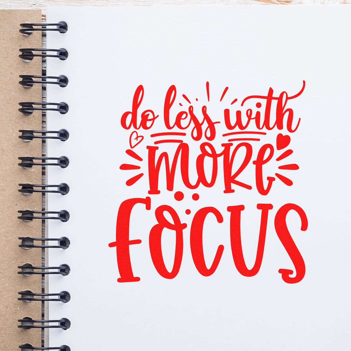 Red stamp seal reading do less with more focus on a white spiral notebook with black rings and kraft cover.