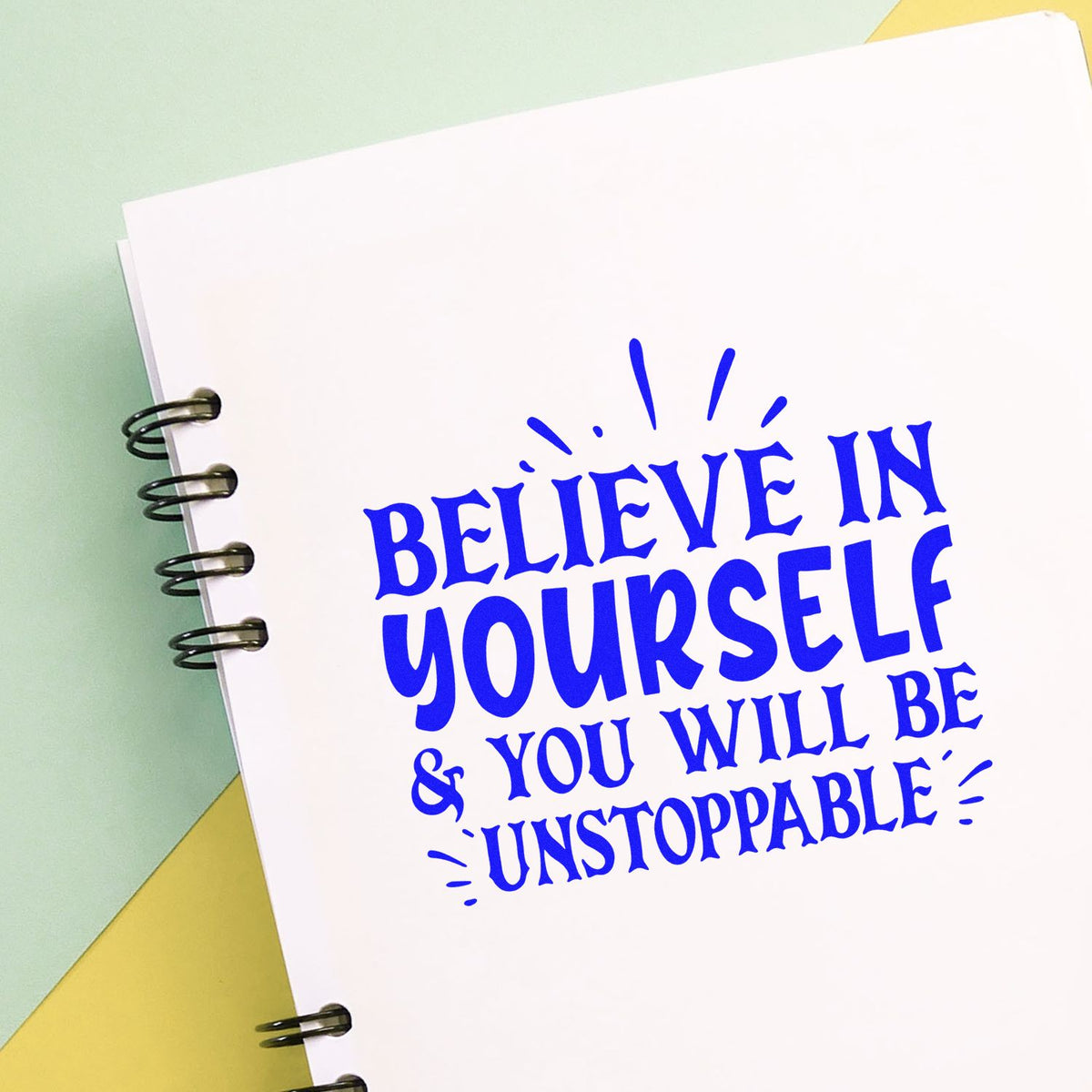 Blue motivational stamp and seal on white spiral notebook, reading Believe in yourself & you will be unstoppable, on pastel background.