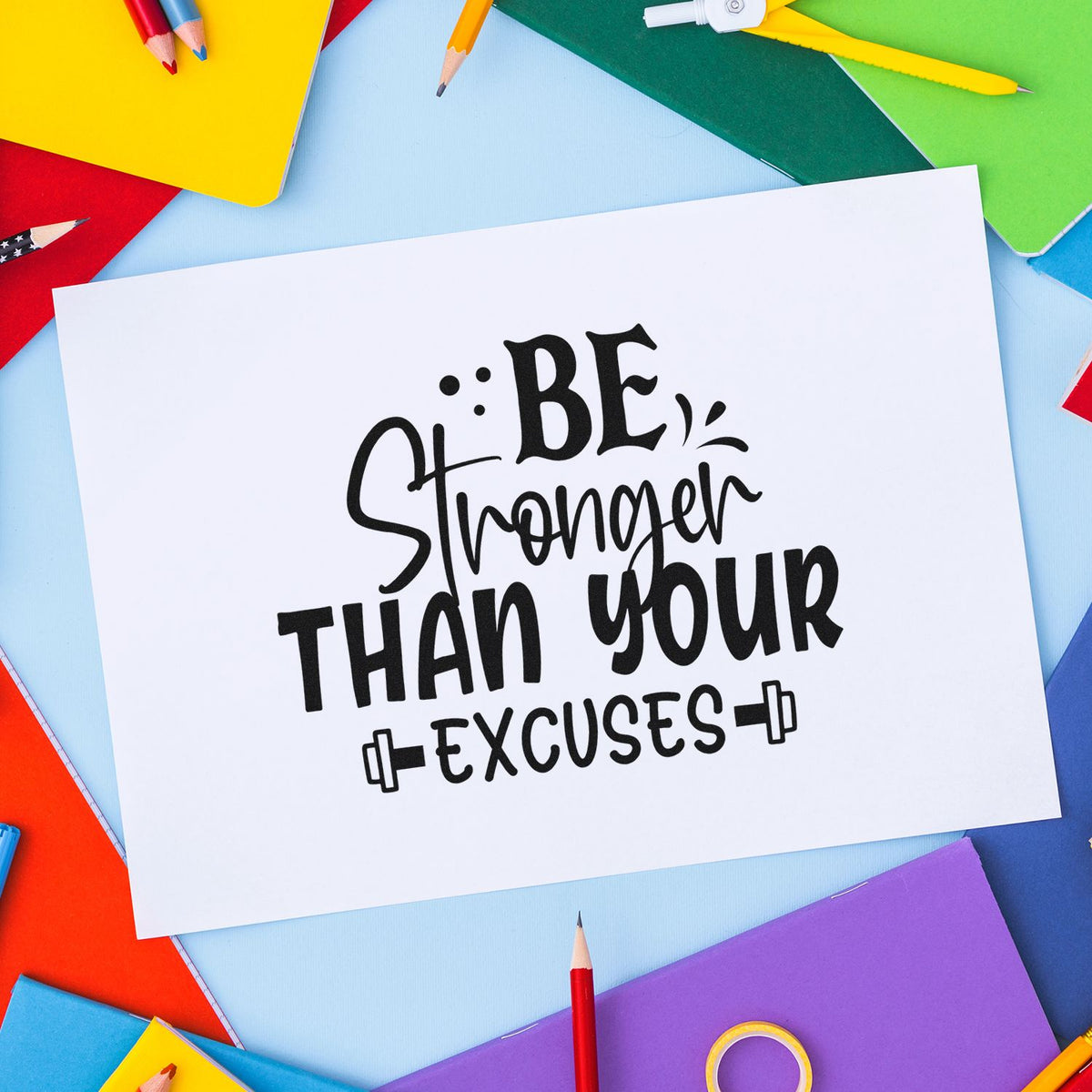 Black custom stamp and seal with Be Stronger Than Your Excuses on white paper, surrounded by colorful pencils and notebooks.