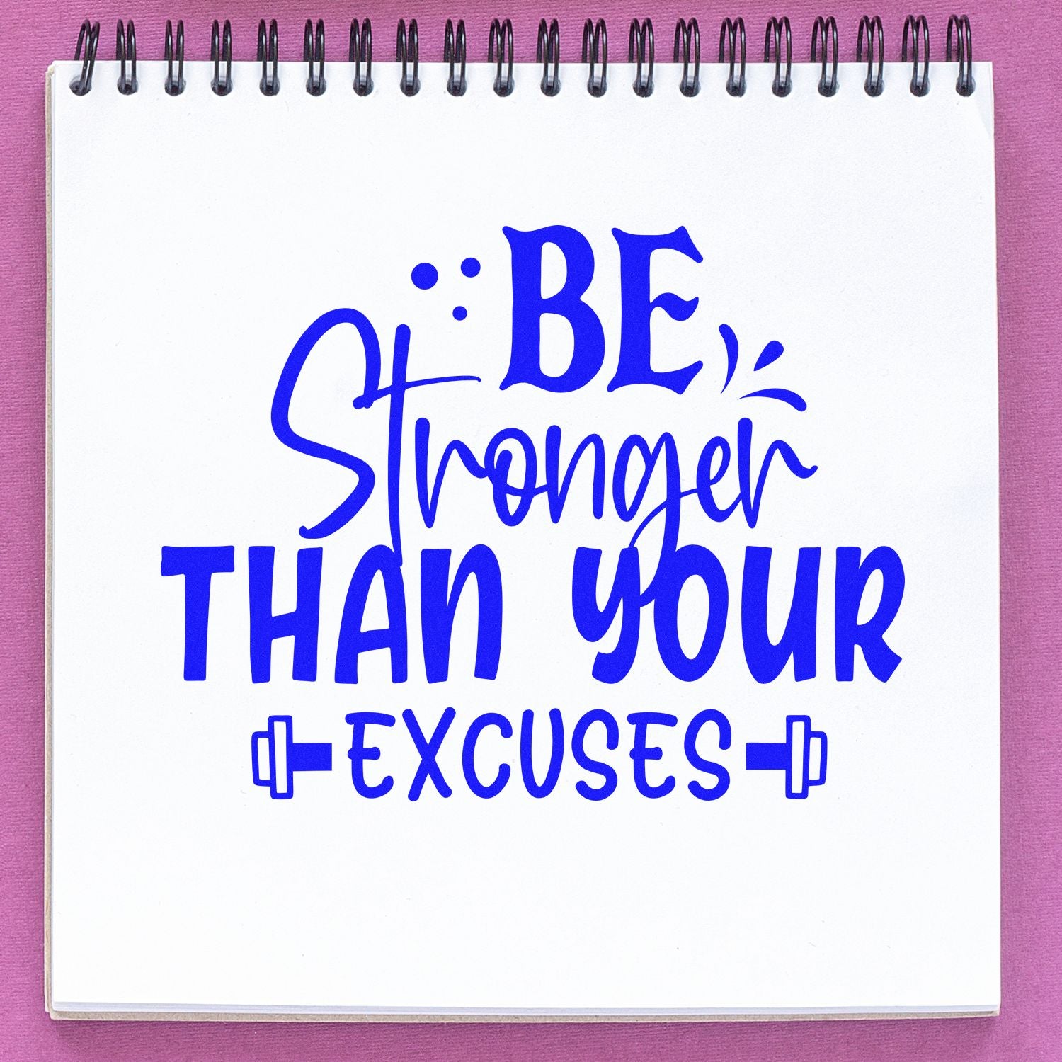 Blue stamp and seal design reading Be Stronger Than Your Excuses on a white spiral notebook with pink background.