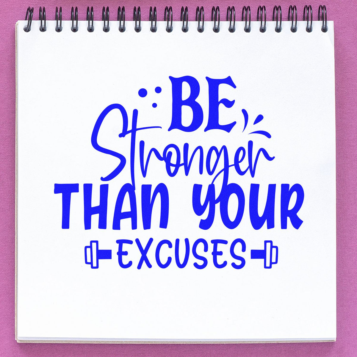 Blue motivational stamp design reading Be Stronger Than Your Excuses on a white spiral notebook with pink background