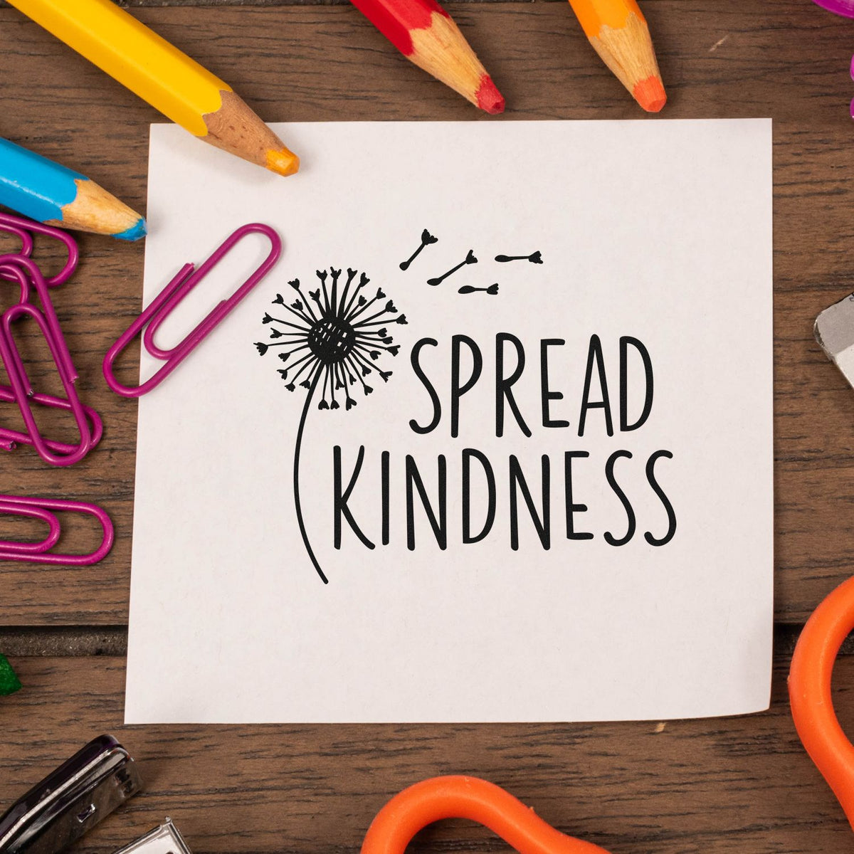 Black rubber stamp reading Spread Kindness with dandelion and seeds on white paper, framed by colorful pencils and purple paperclips.