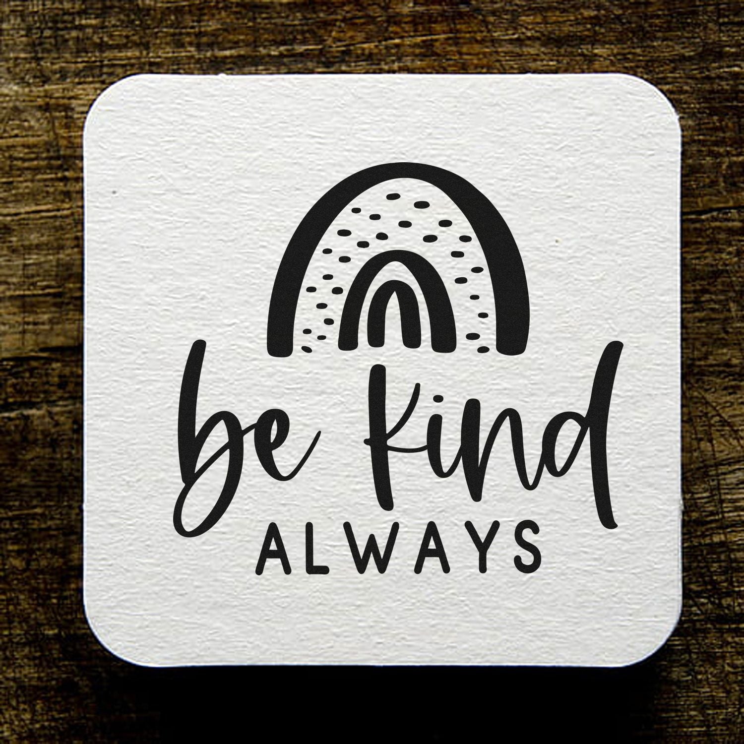 White textured square stamp seal with black rainbow and be kind ALWAYS text on rustic dark wood background.