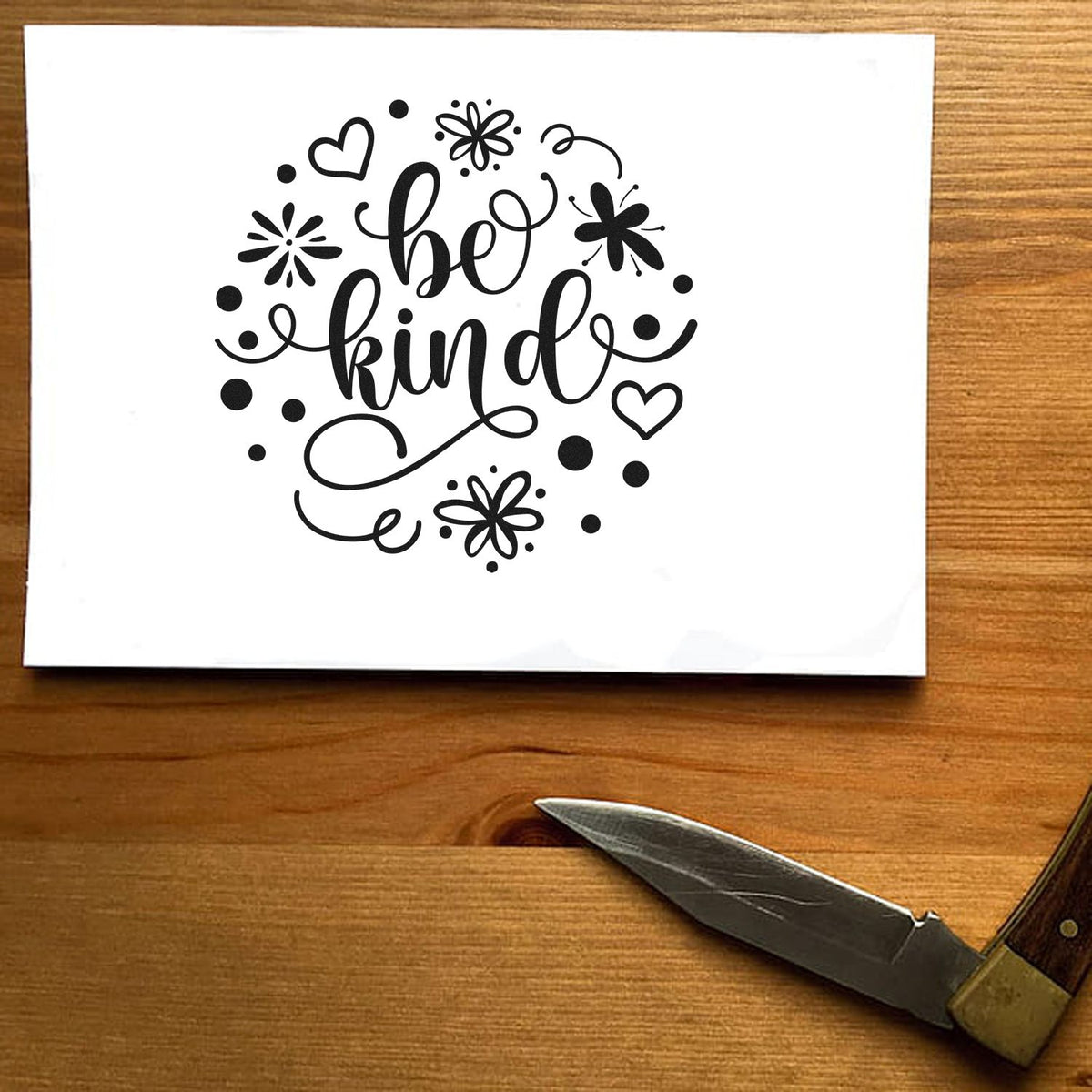 Black Be Kind rubber stamp design with hearts, flowers, and dots on a white card, on a wooden table with carving knife.