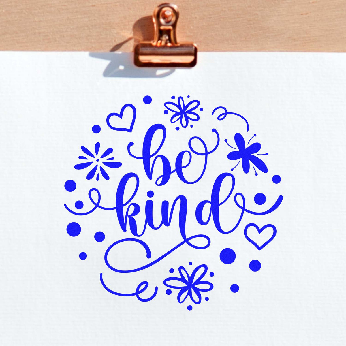 Blue be kind decorative stamp seal with hearts, flowers and dots, clipped to white paper with a copper binder clip.