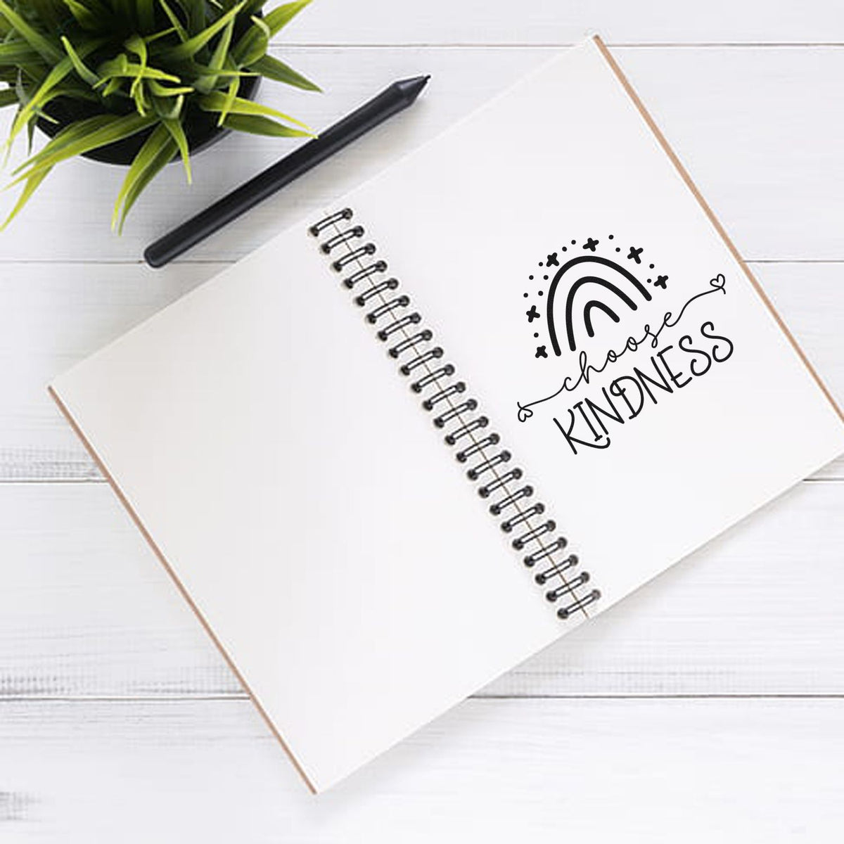 White spiral notebook with black choose kindness rubber stamp design, black pen, and green plant on a white desk.